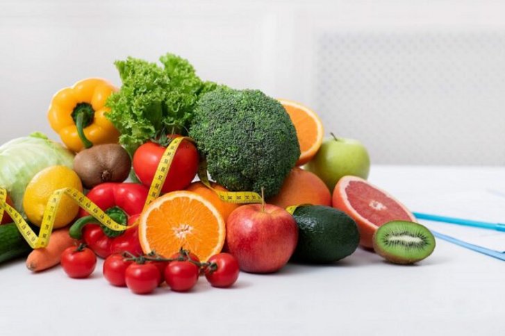 Stilleben zum Thema Ernaerungsberatung auf dem ein grosser Haufen buntes Obst und Gemuese, ein Glas Saft, ein Massband und ein Klemmbrett zu sehen ist.